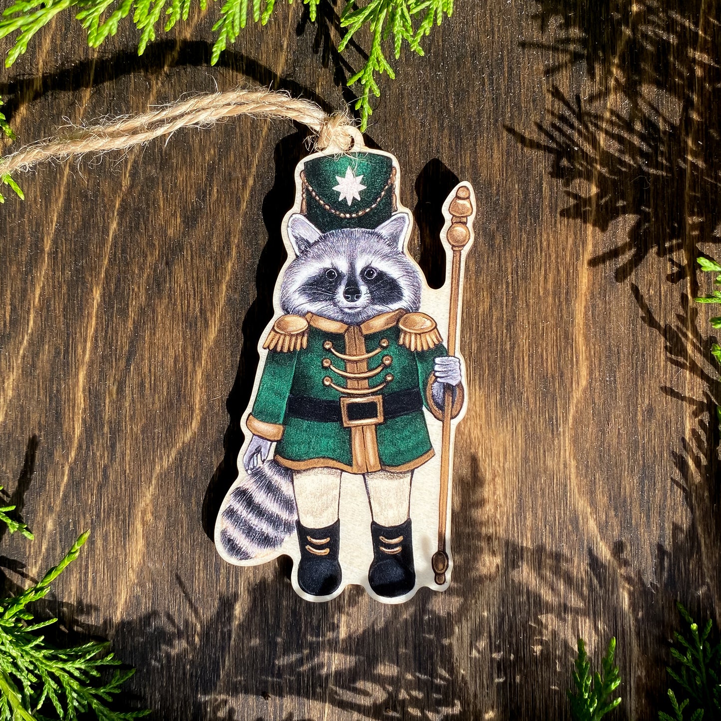 A wooden ornament strung with twine of a raccoon in a green nutcracker outfit.