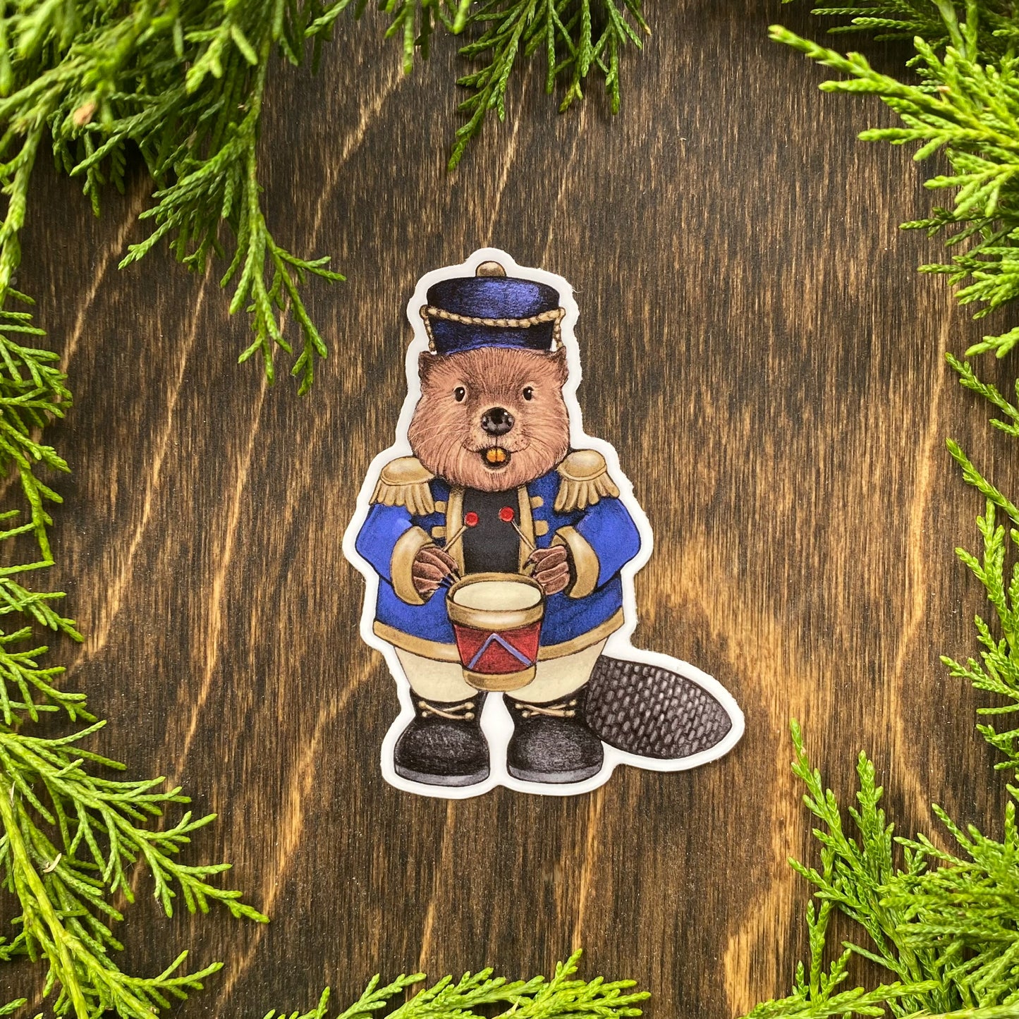 A weatherproof vinyl sticker of a beaver dressed in a blue nutcracker outfit surrounded by cedar foliage.