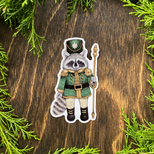 A weatherproof vinyl sticker of a raccoon dressed in a green nutcracker outfit surrounded by cedar foliage.