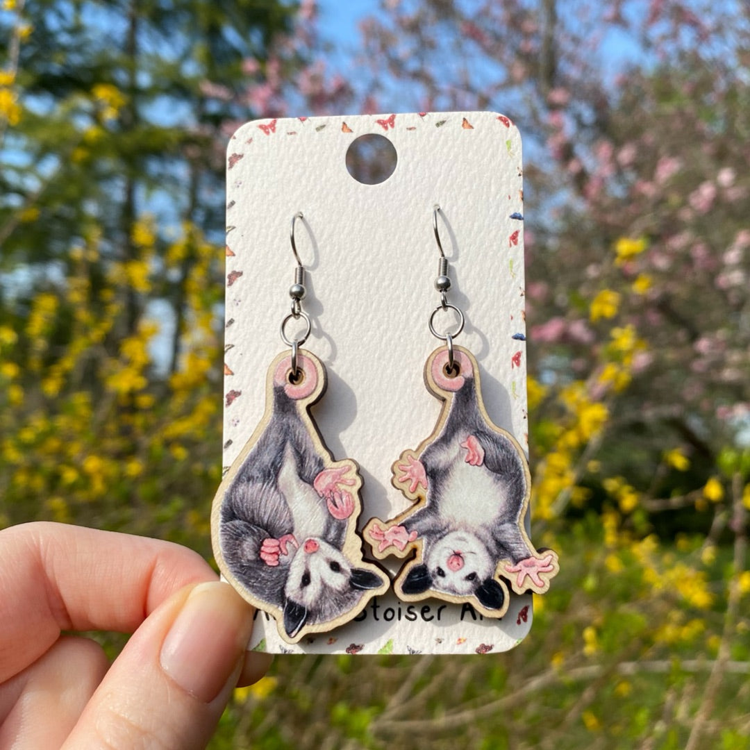 A hand holding wooden earrings of baby opossums hanging upside down.