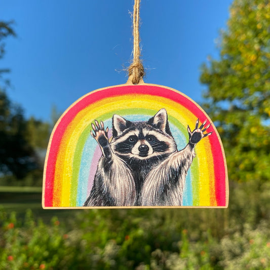 A wooden ornament strung with twine of a raccoon with its hands raised in front of a rainbow.