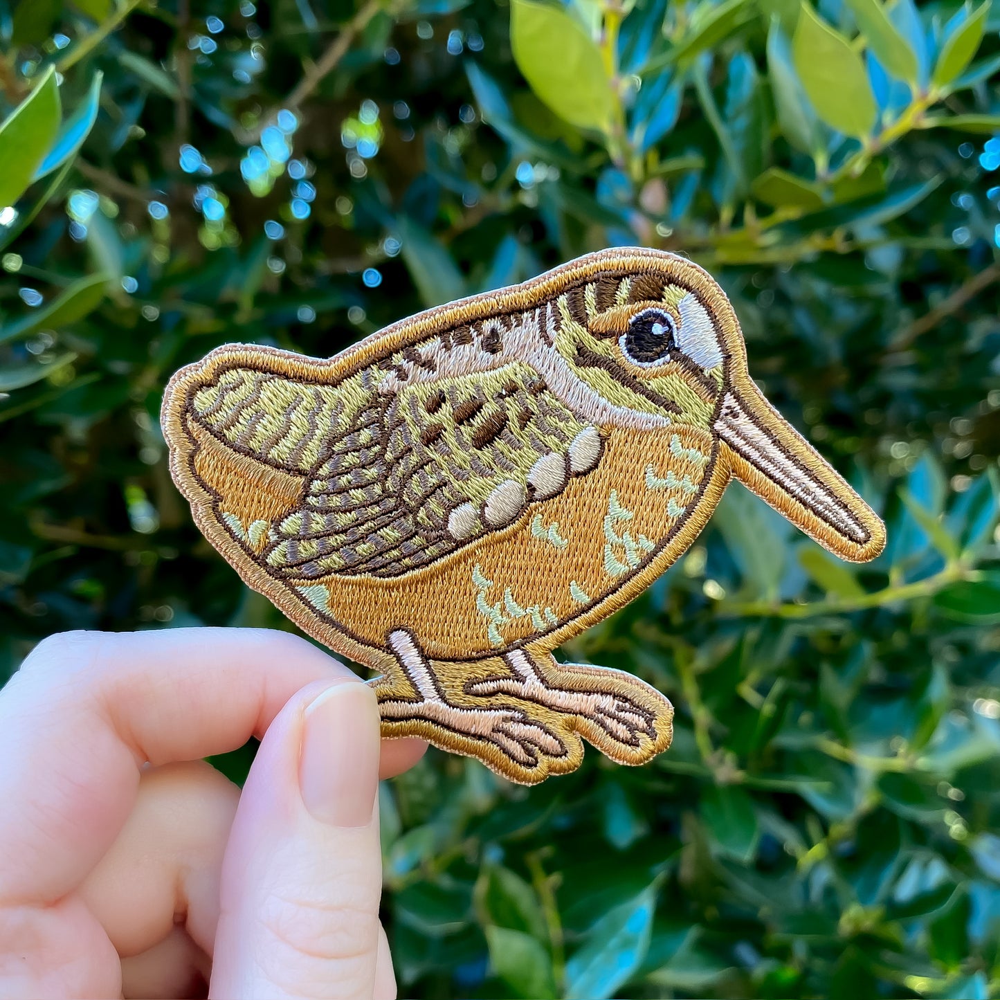 An embroidered patch of an American woodcock held against a blurred background of greenery.