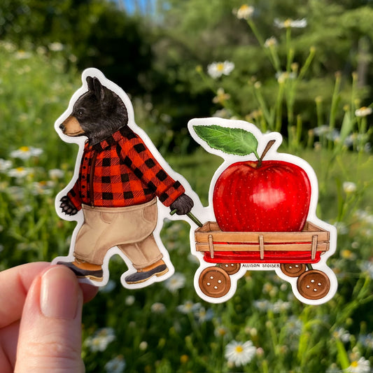 Sticker of a bear in a red plaid jacket pulling a cart with an apple, held against a natural background.