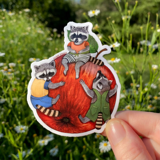 Sticker of three raccoons in colorful outfits on a red apple held in front of a natural background with greenery and flowers.