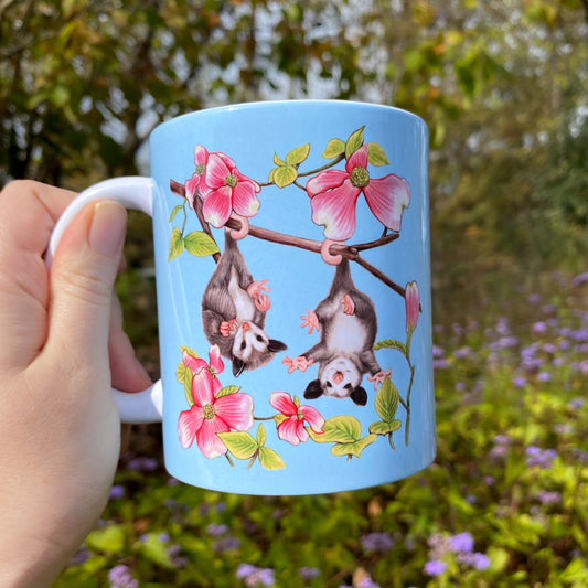 Light blue mug with an opossum and dogwood illustration held by a hand against a natural background