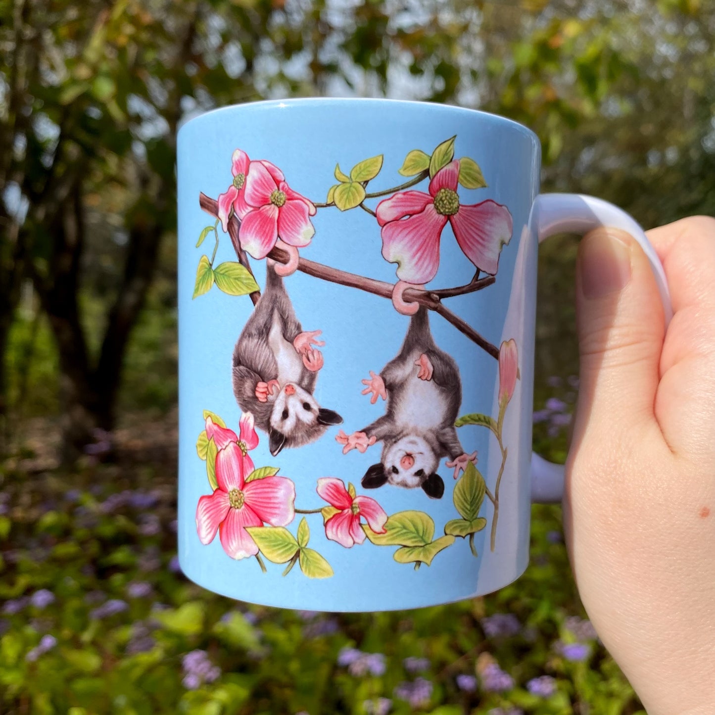 Light blue mug with a baby opossum and dogwood illustration held in a hand outdoors.