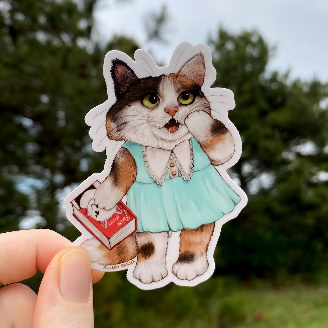 Sticker of a calico cat in a blue dress held by a hand with a blurred natural background