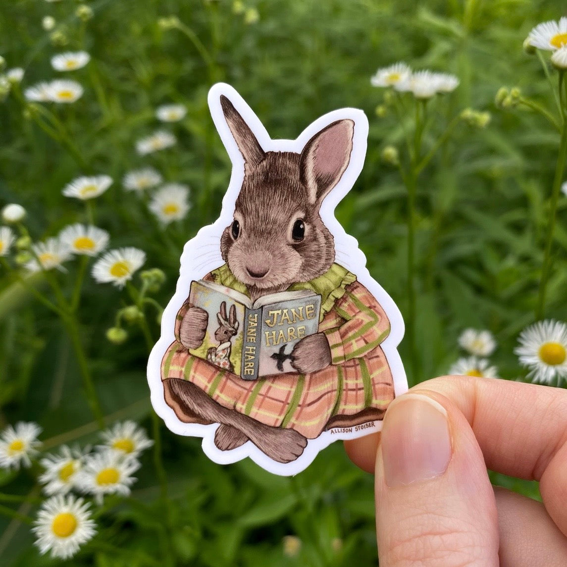 Sticker of a rabbit holding a book in a field with flowers. The book's title is Jane Hare.