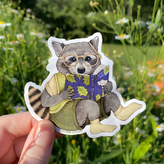 Sticker of a raccoon wearing glasses and reading a book held in front of a natural background with flowers and grass.