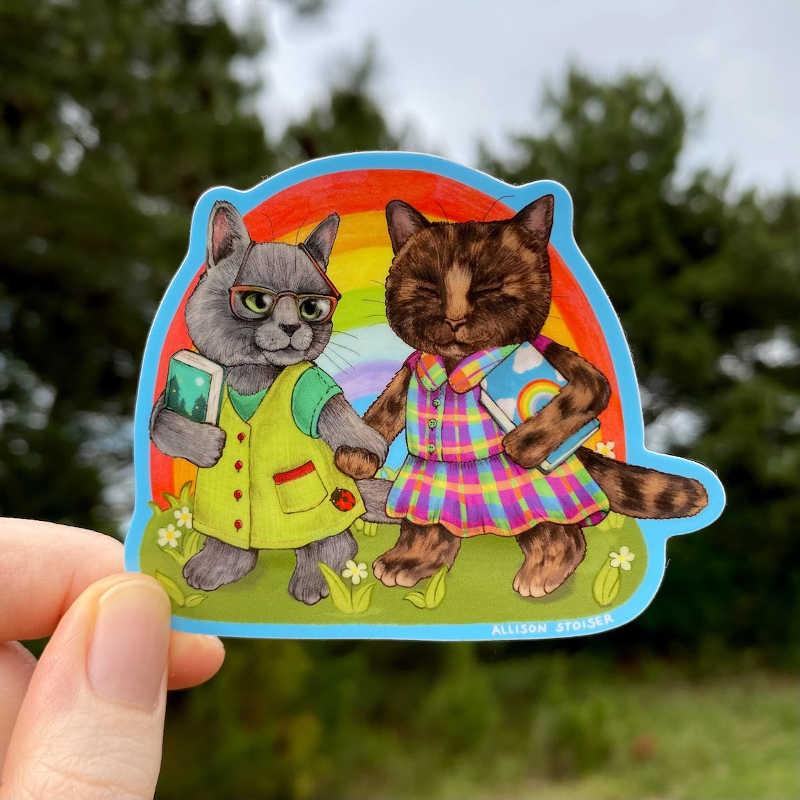 Sticker of two cats holding hands with a rainbow background, held by a hand outdoors.