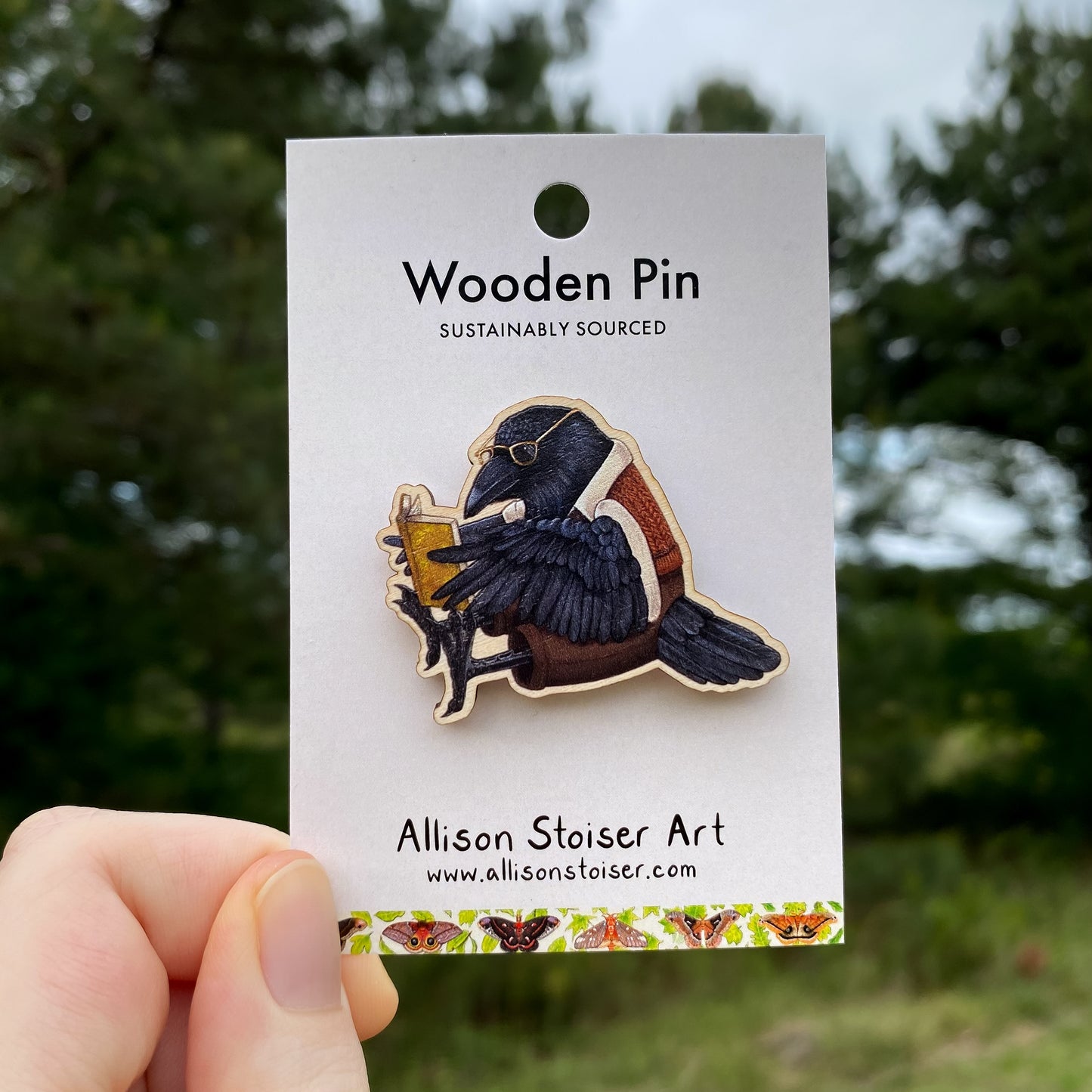 Wooden pin of a raven holding a book, on a card with 'Allison Stoiser Art' branding, against a blurred natural background.