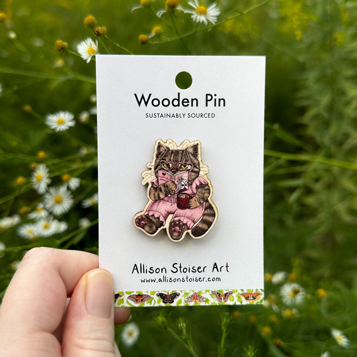 A hand holds a wooden pin on a white backing card outdoors in a field of asters and green foliage. The pin depicts a reading tabby cat in a pink dress holding a small red mug.