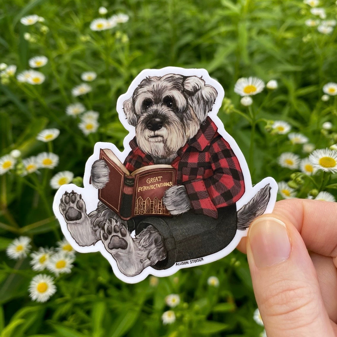 Sticker of a schnauzer in a plaid shirt reading a book held in front of a natural background with flowers.