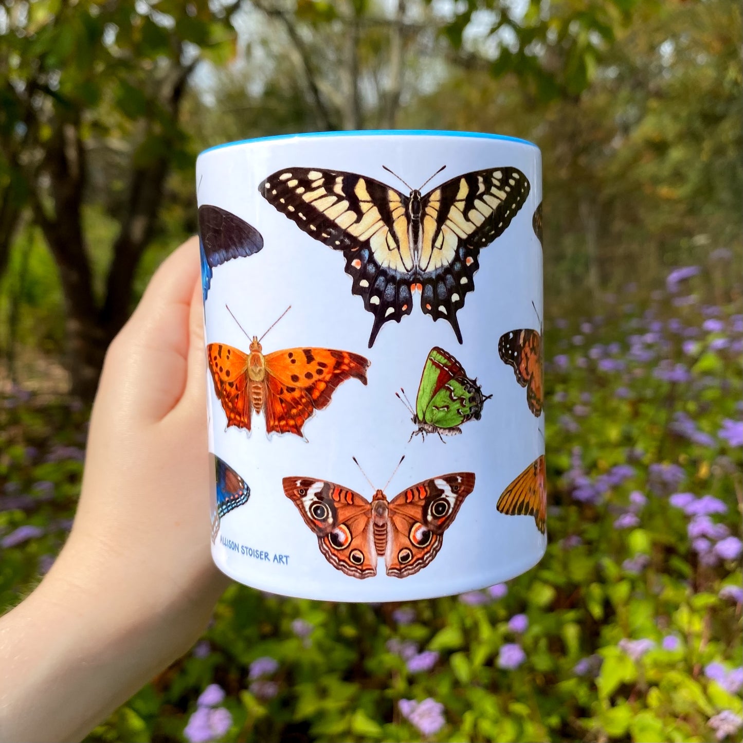 Mug with butterfly illustrations held in front of a natural background with flowers and trees.