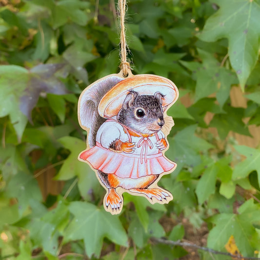 A wooden ornament strung with twine of a Douglas squirrel in a porcini hat and pink skirt.