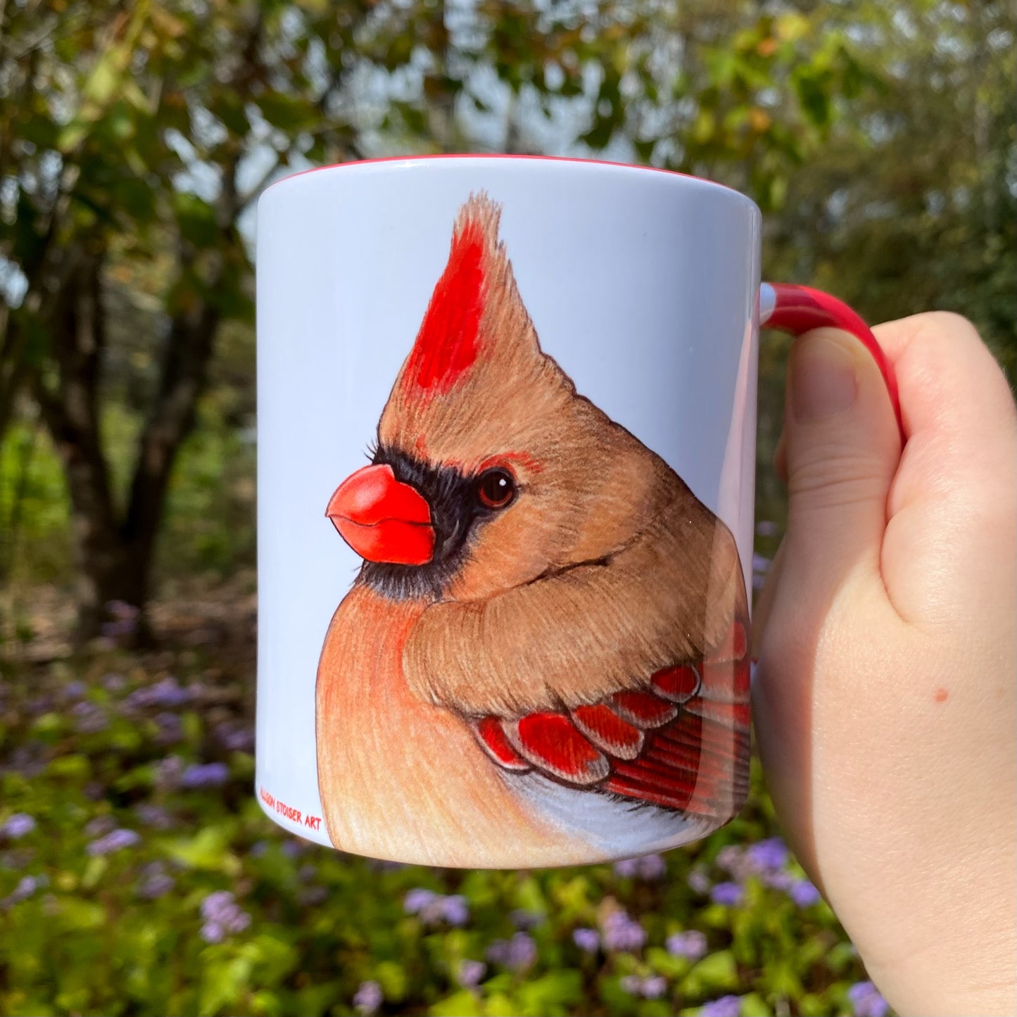 Mug with a female cardinal illustration held by a hand in a natural setting.