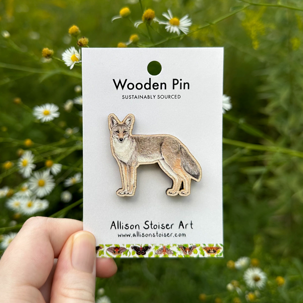A hand holds a white display card featuring a wooden coyote pin. The card reads 'Wooden Pin' and 'Allison Stoiser Art' with the website allisonstoiser.