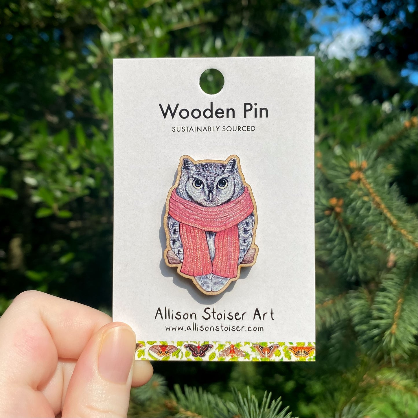 Wooden pin with a screech owl in a pink scarf illustration on a card held in front of green foliage