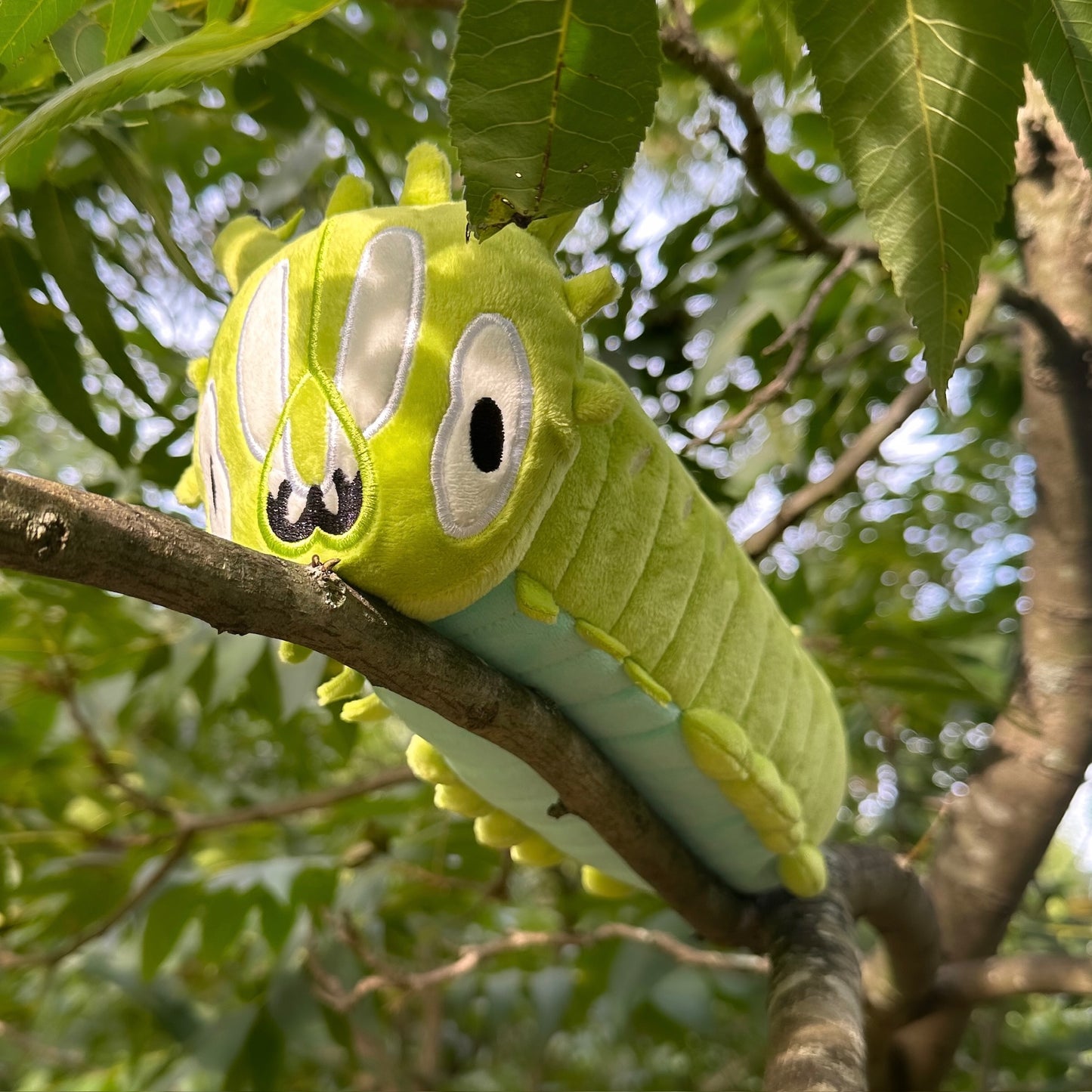 A green caterpillar plush toy with embroidered eyes and mouth, resting on a tree branch outdoors among green leaves.