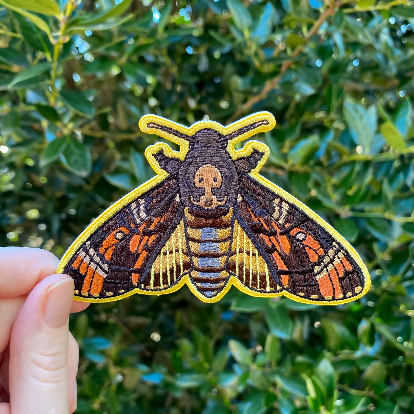 An embroidered patch of a death's-head hawkmoth held against a blurred greenery background.
