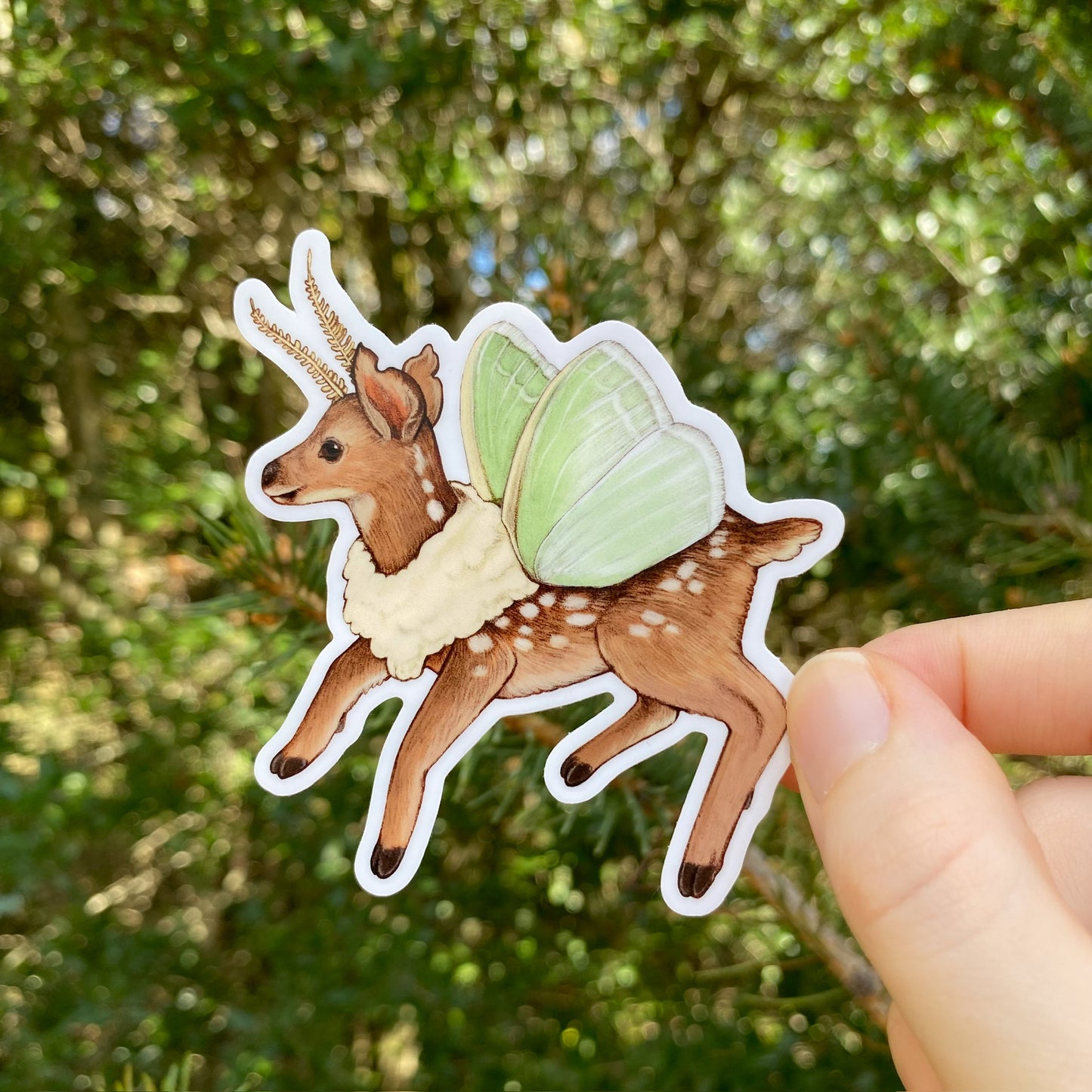 A hand holding a sticker of a white-tailed deer fawn with emerald moth wings.