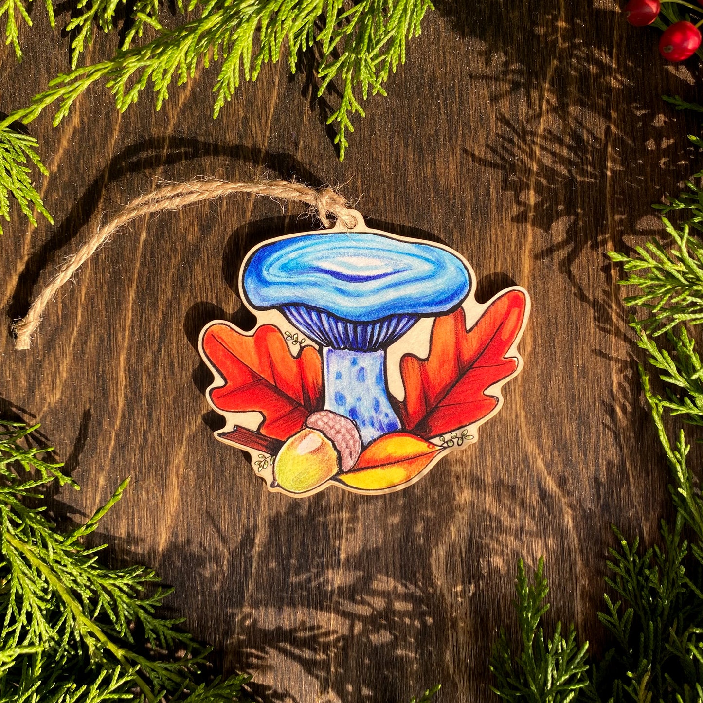 A wooden ornament strung with twine of an indigo milkcap mushroom surrounded by oak leaves.