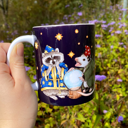 Mug with a wizard raccoon and opossum illustration held by a hand against a natural background