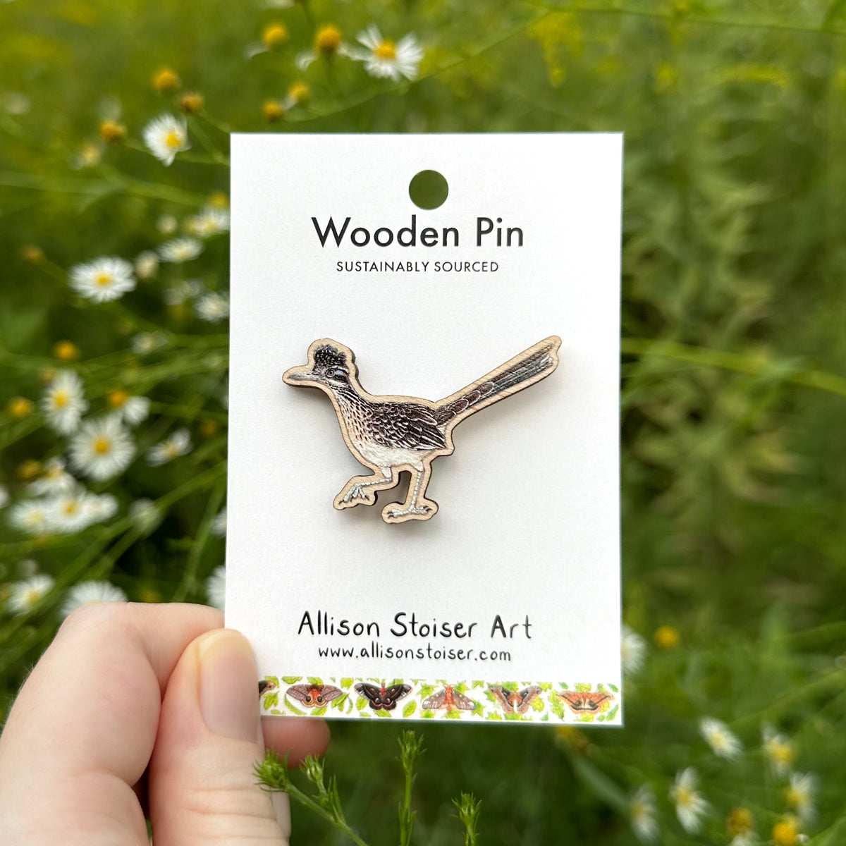 A wooden roadrunner pin displayed on a white backing card held by a hand, with a blurred outdoor background of green foliage and small white flowers.