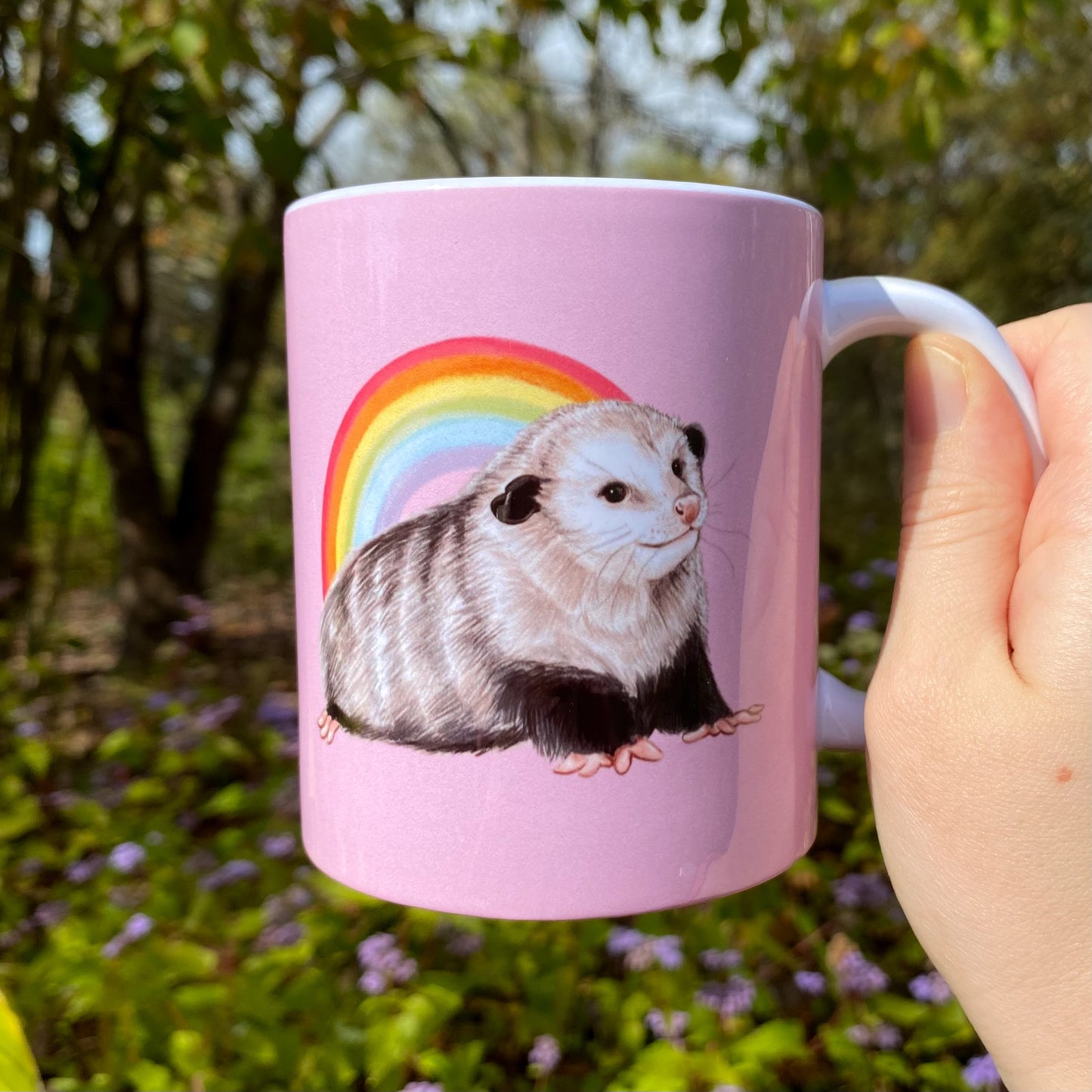 Pink mug with an opossum and rainbow design held in a hand outdoors.