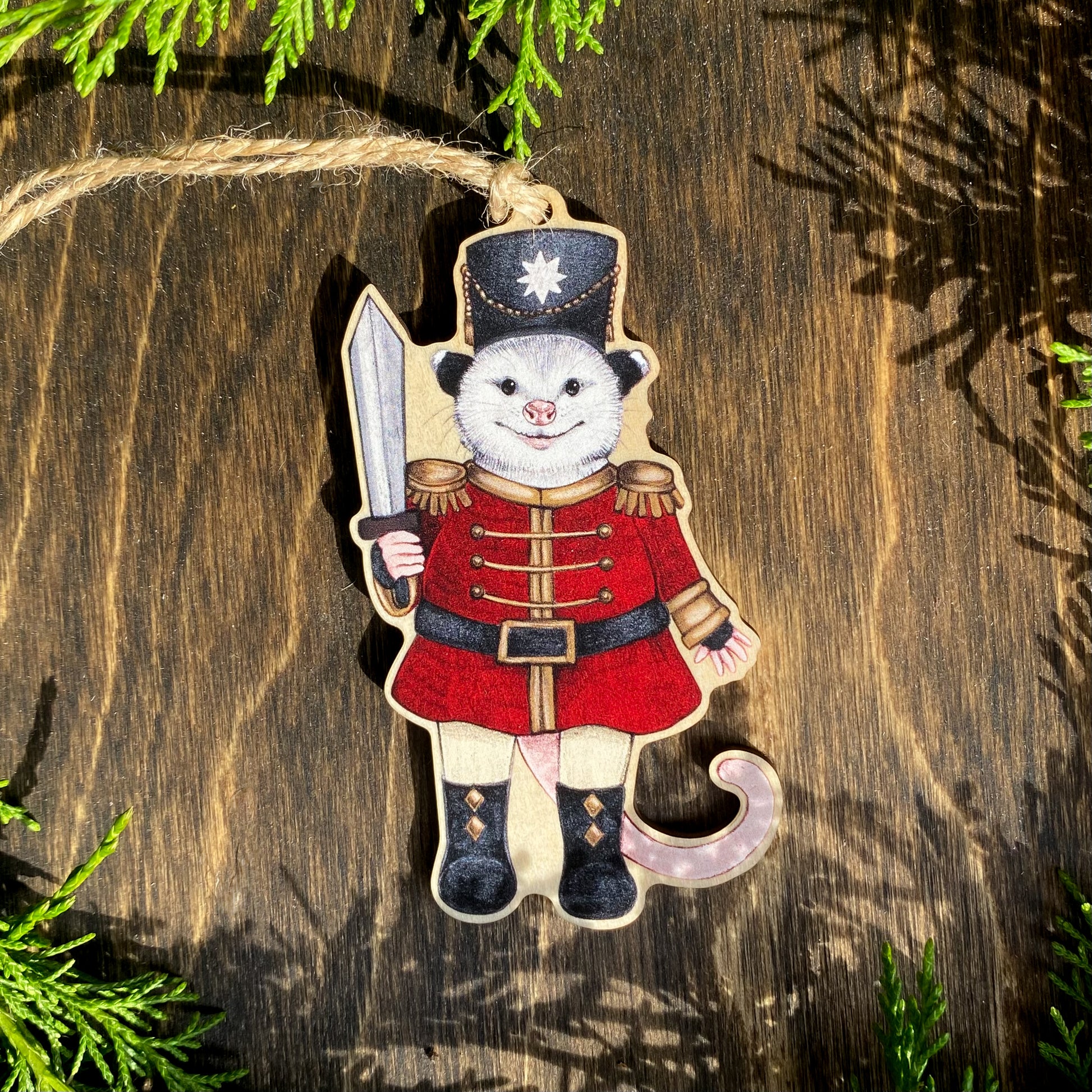 A wooden ornament strung with twine of an opossum in a red nutcracker outfit.