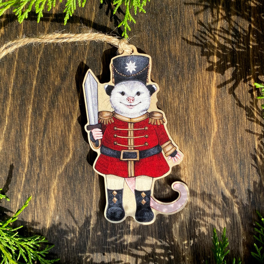 A wooden ornament strung with twine of an opossum in a red nutcracker outfit.