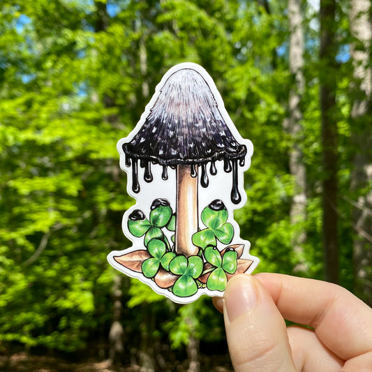 A hand holding a sticker of an inky cap mushroom dripping ink on some clover