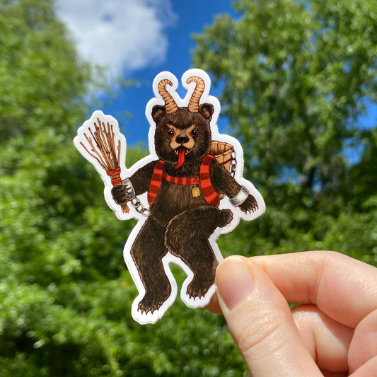 A hand holding a vinyl sticker of a bear dressed as Krampus