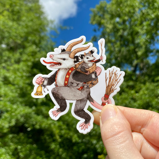 A hand holding a sticker of an opossum dressed as Krampus