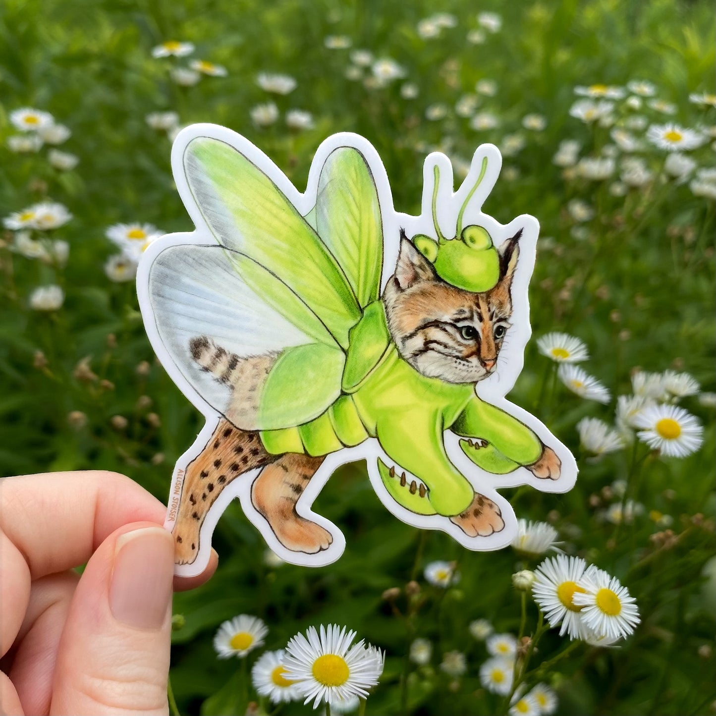 Sticker of a cat in a green praying mantis costume held in front of a grassy background with flowers.