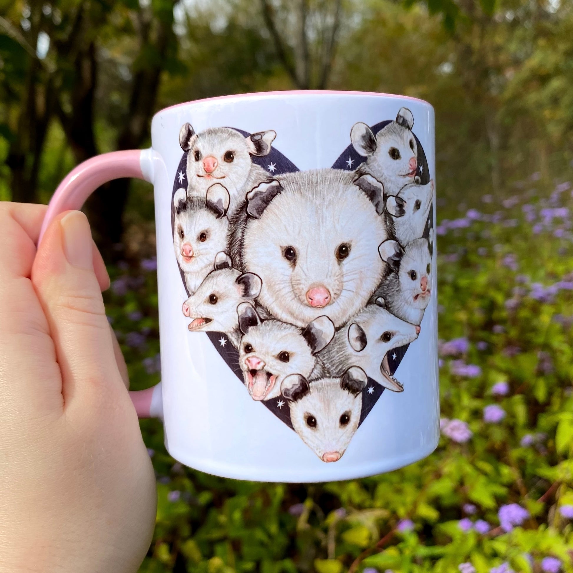 Mug with opossum illustrations held by a hand in a natural setting