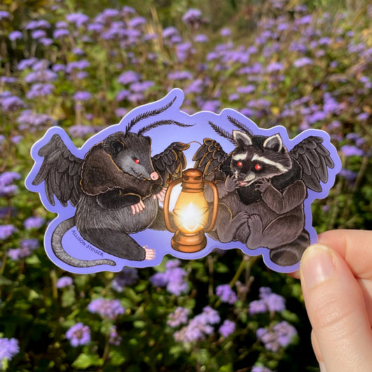 A hand holding a purple sticker of a mothman opossum and mothman raccoon sitting in front of a lantern.