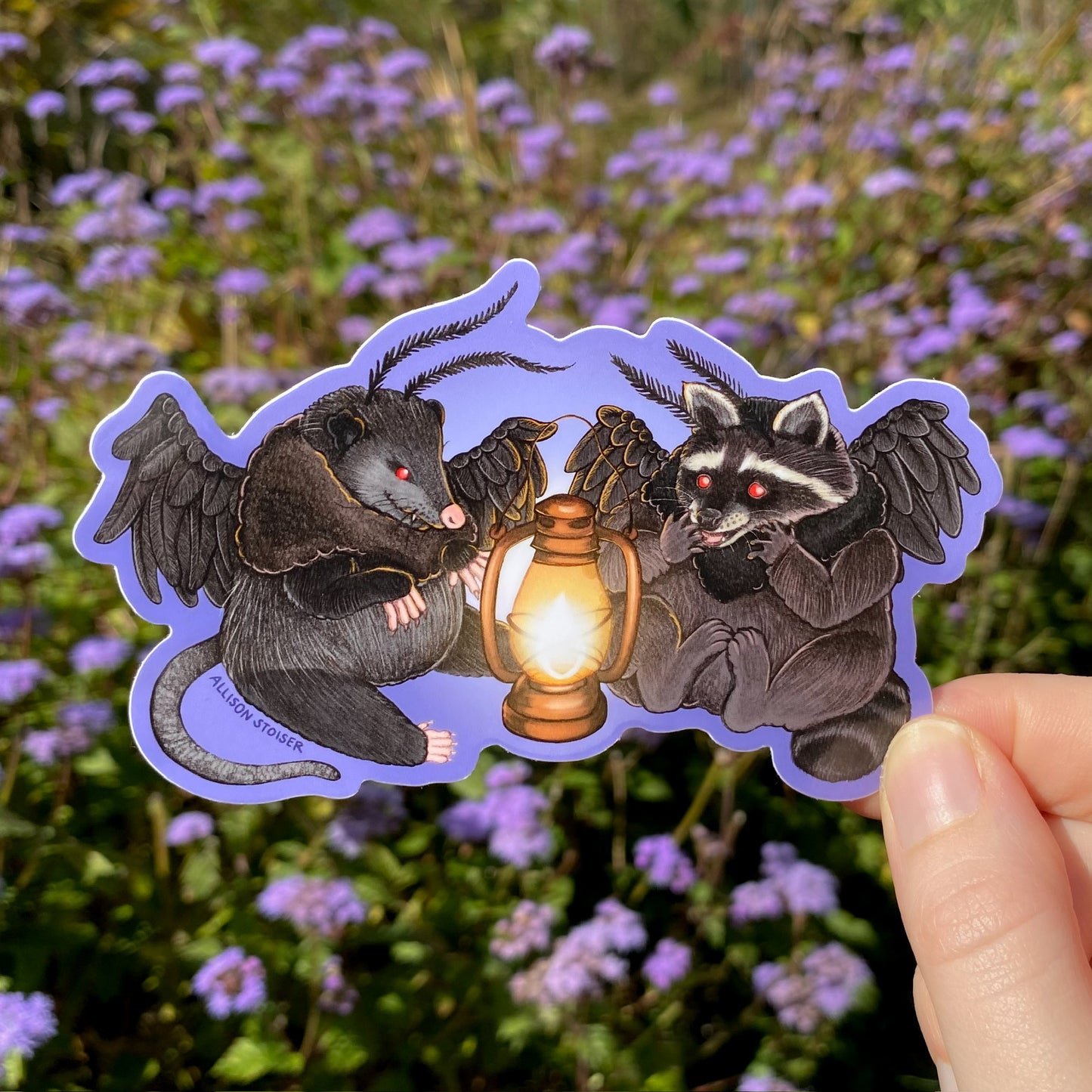 A hand holding a purple sticker of a mothman opossum and mothman raccoon sitting in front of a lantern.