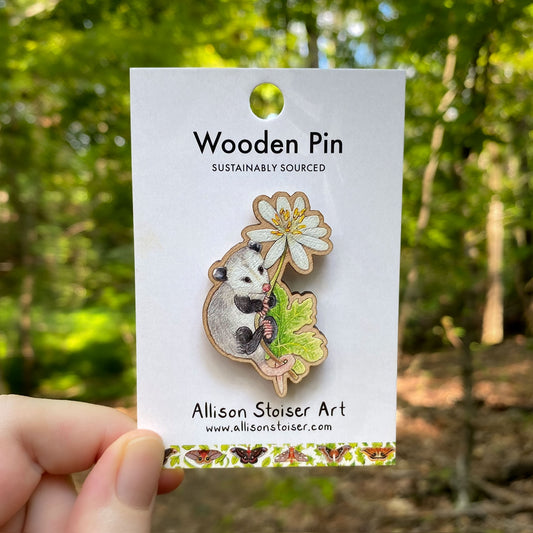 Wooden pin with an opossum clinging onto a stem of bloodroot on a card labeled 'Allison Stoiser Art' against a natural background.