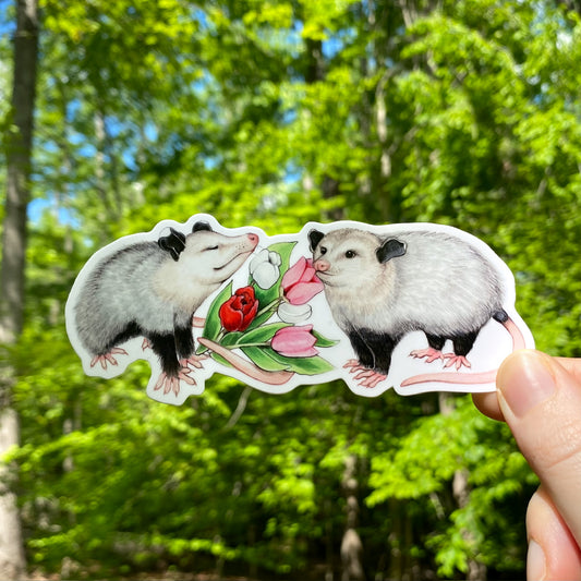 A hand holding a sticker of an opossum presenting a bouquet of tulips to another opossum