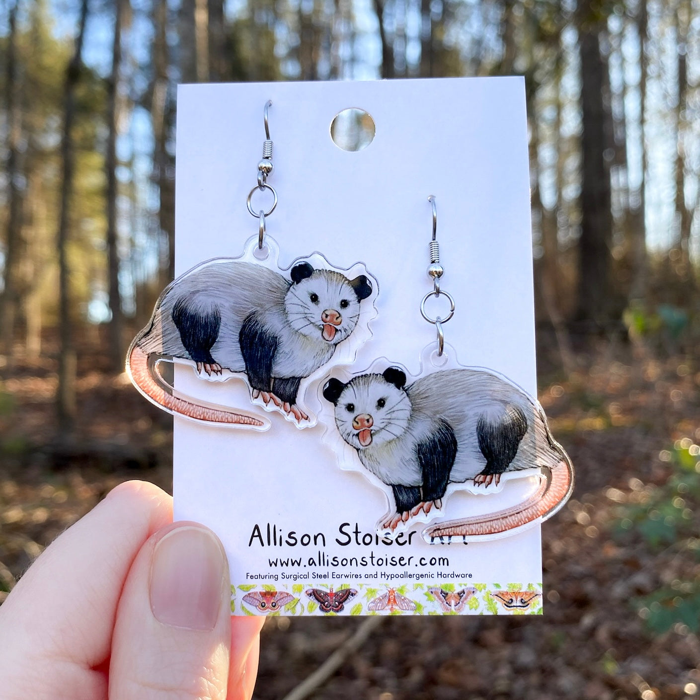 Opossum earrings on a card held in front of a forest background