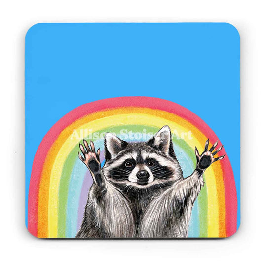 A blue coaster of a raccoon with its arms raised in front of a rainbow on a white background.