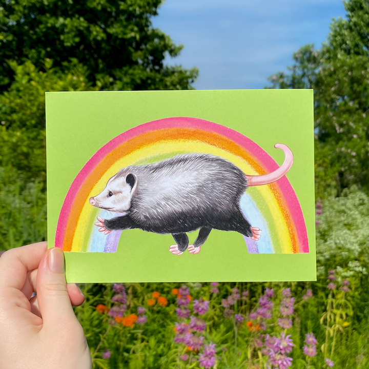 Hand holding a card with an opossum and rainbow illustration against a natural background.
