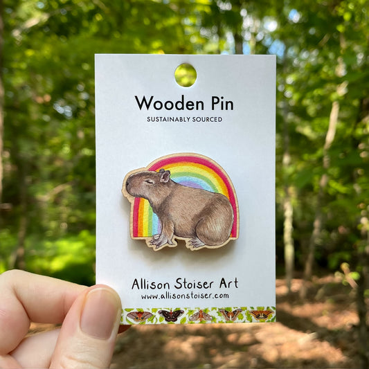 Wooden pin with capybara in front of a rainbow on a card with 'Allison Stoiser Art' branding, held in front of a forest background.