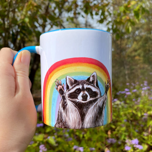 Mug with a raccoon illustration in front of a rainbow held by a hand outdoors.