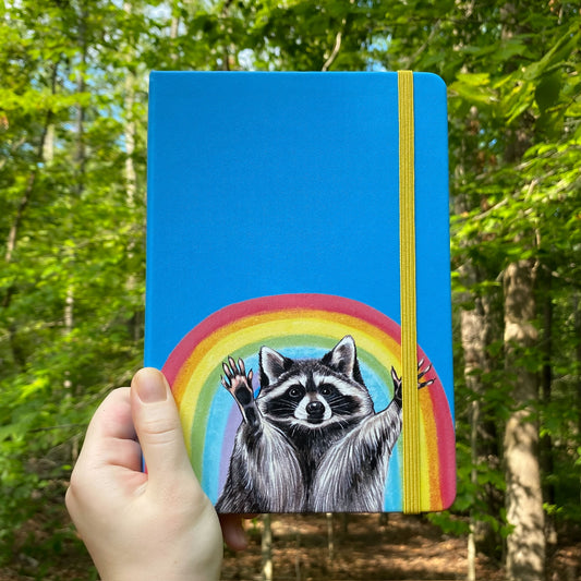 Blue notebook with a raccoon and rainbow held in front of a forest