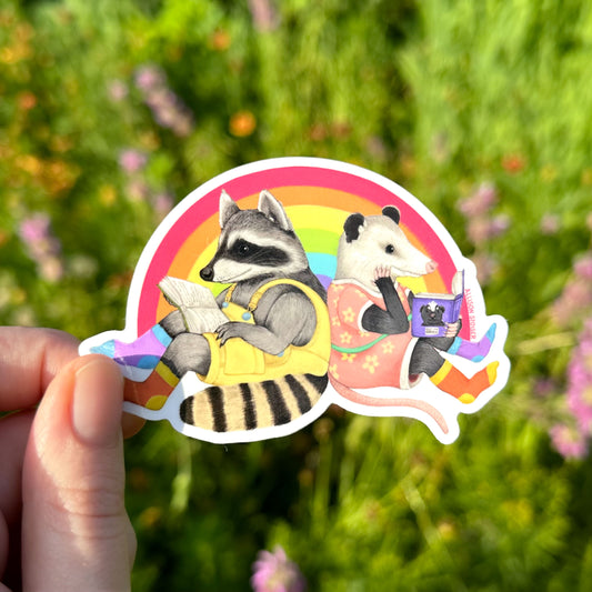 a hand holding a sticker of a raccoon and opossum sitting back to back and reading in front of a rainbow.