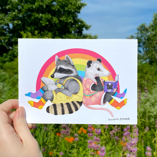 Illustration of a raccoon and opossum reading books with a rainbow background, held in front of a natural background.
