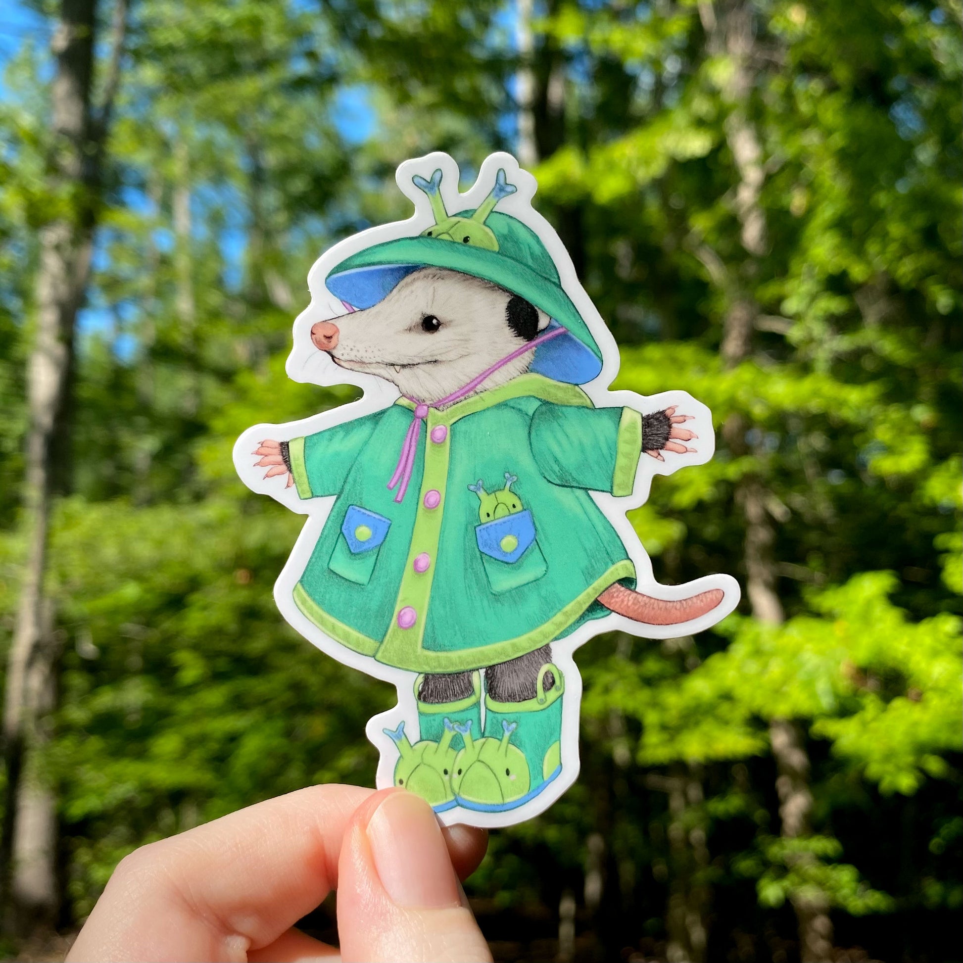 A sticker of an opossum in a blue-green raincoat and rain boots.