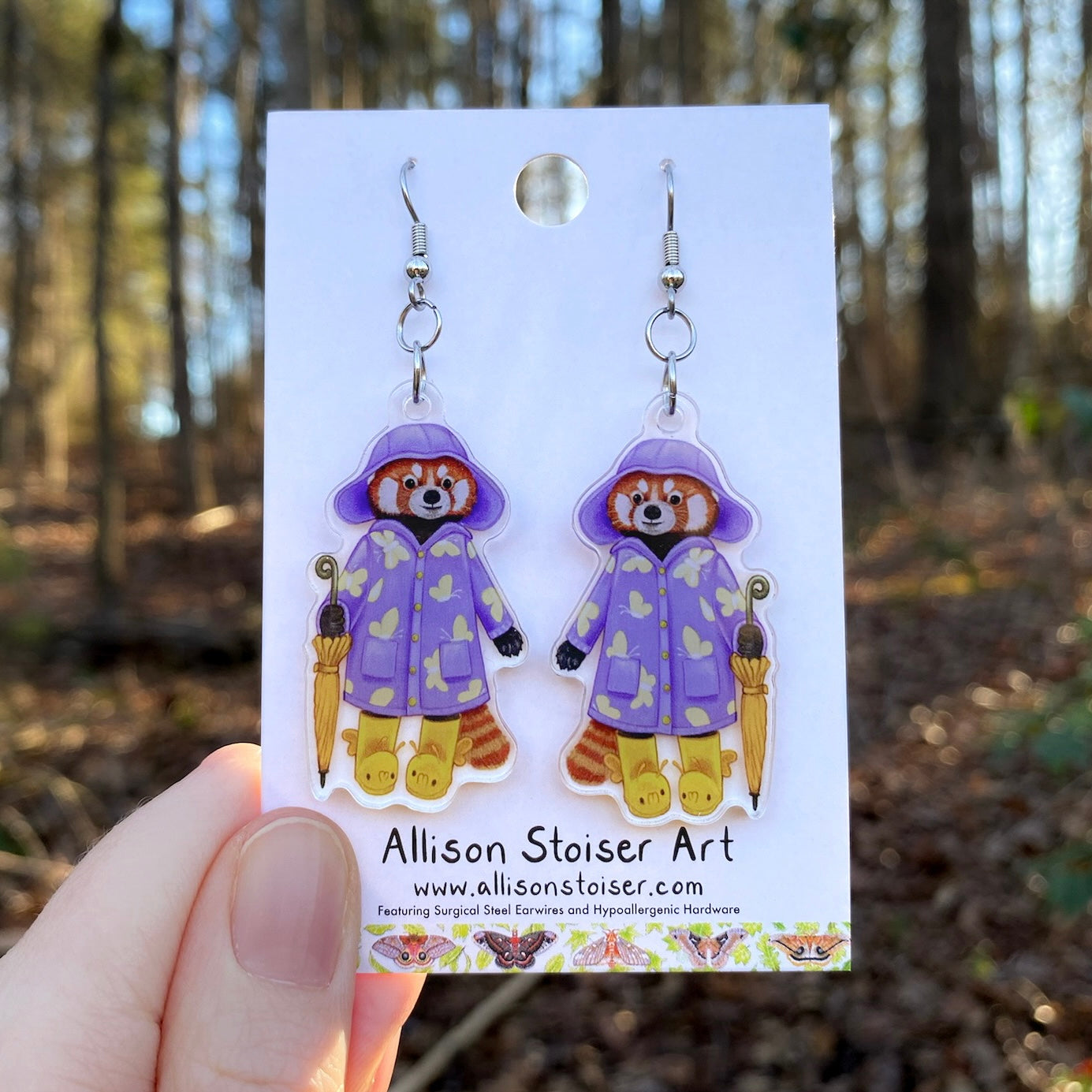 Earrings with red panda design in raincoats held on a card with 'Allison Stoiser Art' branding, against a blurred natural background.
